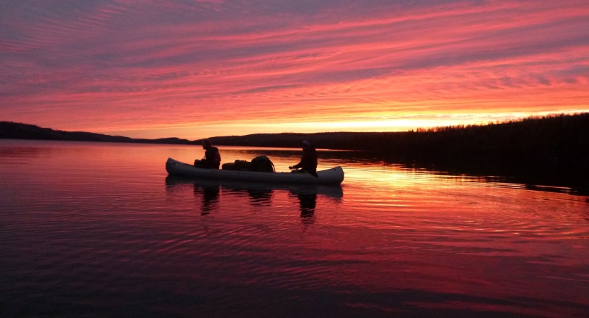 The sky appears in shades of red, orange and purple, reflected in the lake below. On the water, the silhouette of a canoe containing two people can be seen.    